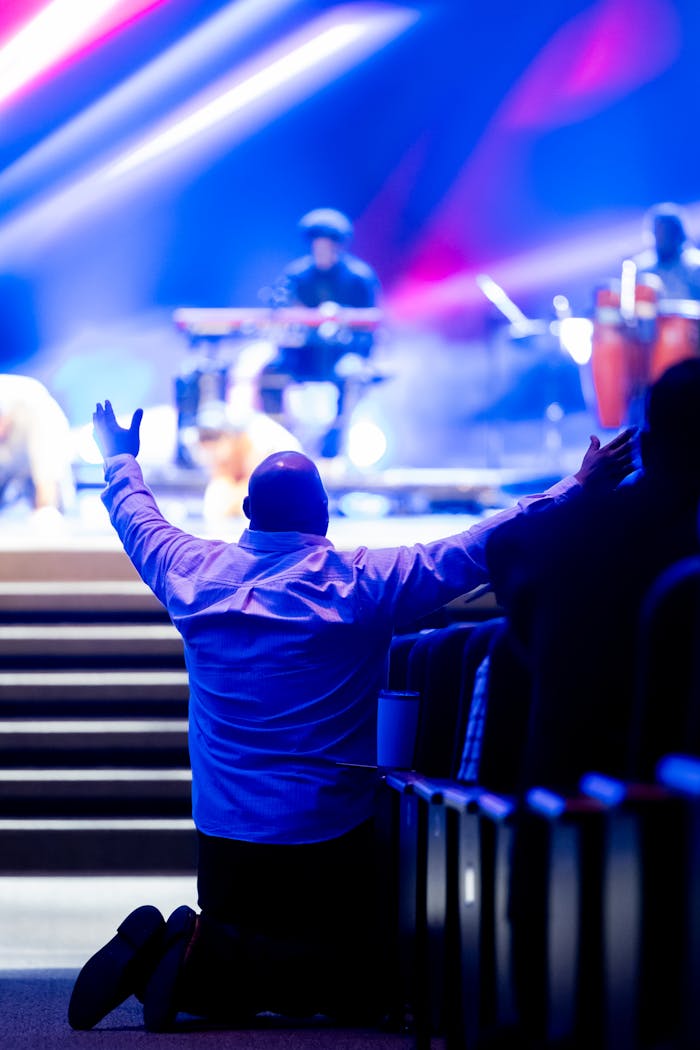 ours-journey A man kneels with arms raised during a live worship concert with vibrant lighting.